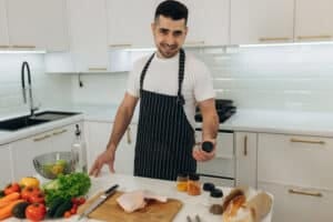 Aaron smiling in a bright kitchen, holding a spice jar beside fresh vegetables and chicken