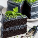 Black Velvet Cake: Simple Scratch Recipe with Black Frosting 3 A slice of dark chocolate cake with black frosting, garnished with green mint leaves and green sprinkles, sits on a white plate with a fork. The whole cake, similarly decorated, is in the background.