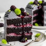 Blackberry Velvet Cake: Easy Gothic Inspired Dessert 3 A slice of dark chocolate layer cake with grey frosting, blackberry filling, and whole blackberries on top, garnished with small green flowers, sits on a white plate with a fork.