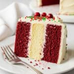 Easy Christmas Velvet Cake: Simple Holiday Dessert 3 A slice of festive cake with red velvet and yellow layers, topped with white frosting, red and green sprinkles, and three red berries, sits on a white plate with a fork beside it.