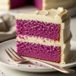 Easy Purple Velvet Layer Cake: Simple Baking 3 A slice of vibrant purple cake with two layers, separated and topped by creamy white frosting, sits on a white plate with a fork beside it. Another part of the cake is visible in the background.