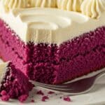 Purple Velvet Cake: Easy White Chocolate Frosting 3 A slice of vibrant purple cake with creamy white frosting on top sits on a plate. A fork holds a bite-sized piece of the moist cake, showing its soft texture and the frosting’s smooth, decorative swirls.